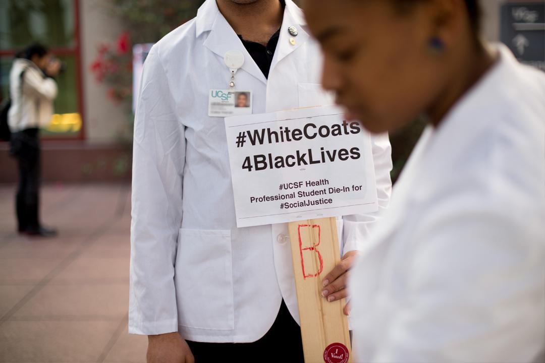 Two UCSF learners wearing their medical practitioner’s white coat at an outdoors public protest event. The person in focus is holding a sign that reads hashtag white coats four Black lives.