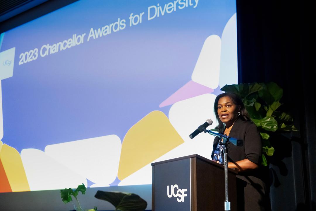 A female presenting speaker with shoulder length hair is giving a speech through a microphone on stage at an indoors UCSF event.