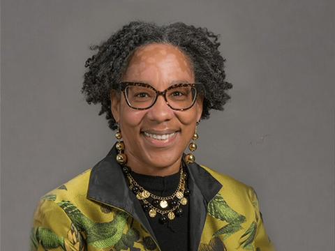 A female-presenting person with short dark hair. They are wearing dark-framed glasses, gold earrings, and a gold necklace. The person is also wearing a light green jacket with floral patterns with a black pointed collar.