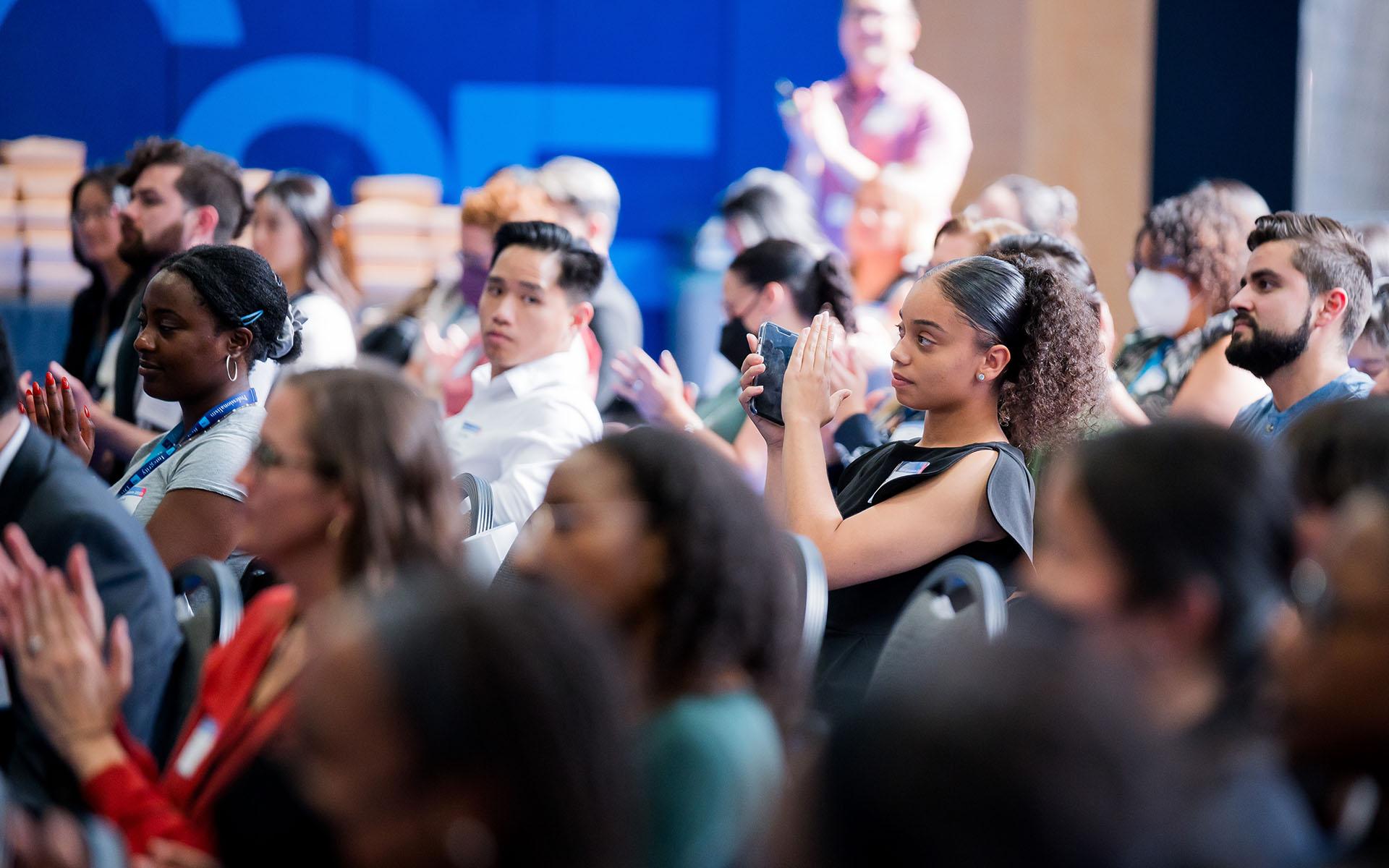 Participants of the 2023 HEAR Symposium are seated in a well-lit gymnasium clapping and watching a speaker at the front of the room out of view.