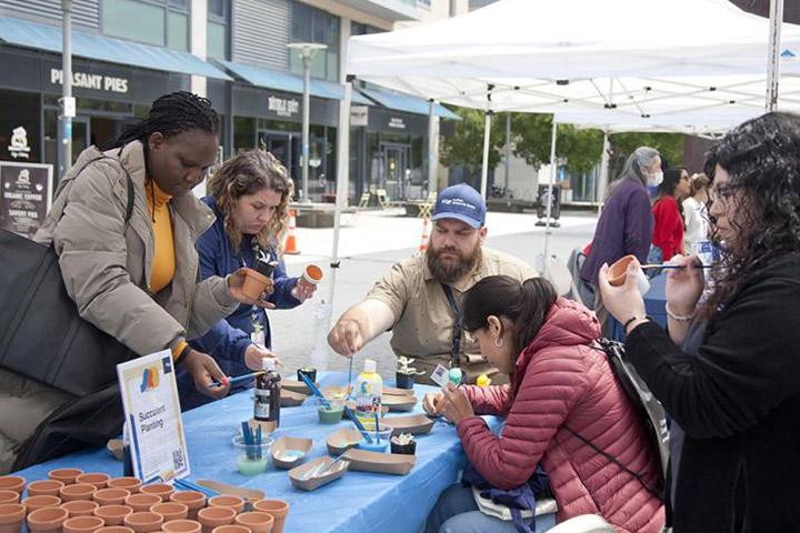 Five UCSF community members attending the 2023 UCSF Staff Resource Day Fair gather around a tented table paint small clay pots they will use to plant succulents they will select.