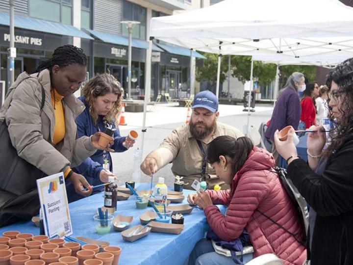 Five UCSF community members attending the 2023 UCSF Staff Resource Day Fair gather around a tented table paint small clay pots they will use to plant succulents they will select.
