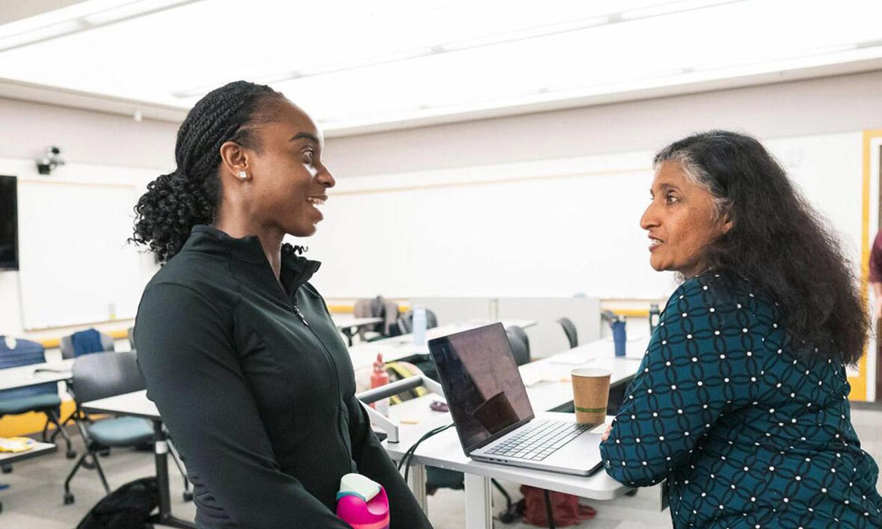 Two women having a conversation at the front desk in an empty classroom.