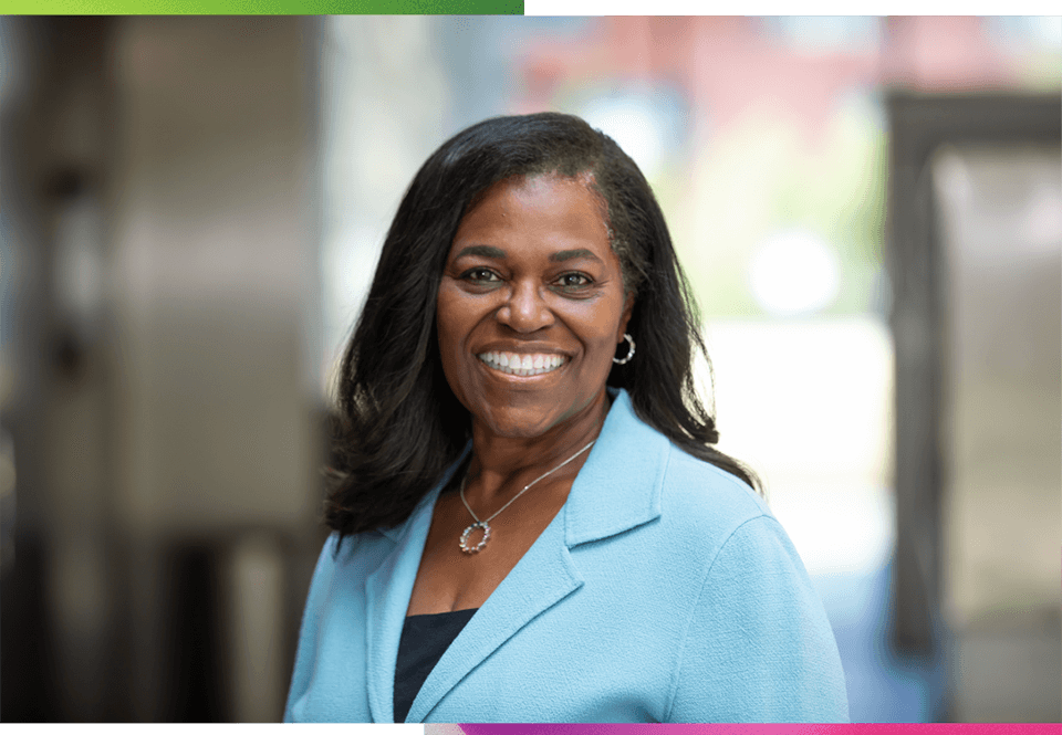 A professional headshot photograph, taken during the day at an indoor lobby office space. A female-presenting person with shoulder-length, long hair, wearing a light blue blazer, facing the camera, and smiling. Graphic elements of gradient pink to purple and green to blues flank the top and bottom corners of the image. A text quote is positioned at the bottom of the image pulled from the Vice Chancellor’s statement.