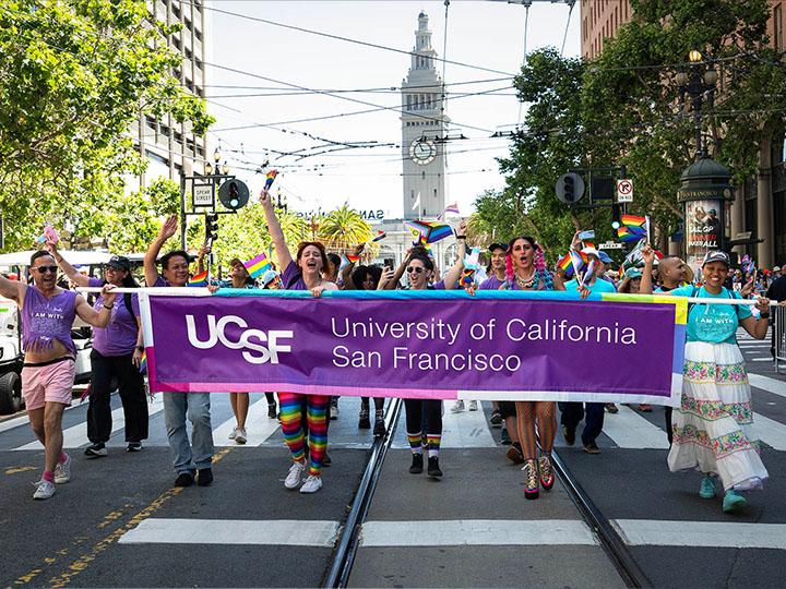 The UCSF community member contingent participates in the 2024 San Francisco Pride parade, proceeding down tree-lined Market Street behind a UCSF branded banner with buildings the Ferry Terminal clock tower in the distance behind them.