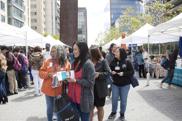 UCSF staff and community members stand in small groups as they make their way down an outdoor open space lined with an aisle of white tents, looking at offerings from each vendor.