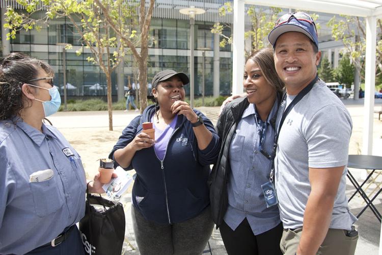 Four UCSF staff members in uniforms chatting and laughing at the Gene Friend Way Mission Bay Campus’s outdoor gathering space.