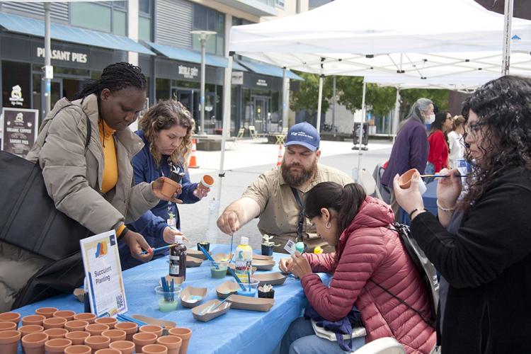 Five UCSF community members gather at an outdoor tent, to paint their own terracotta pots before planting it with a succulent plant. This craft activity occurred during the 2023 Staff Resource Day event at the UCSF Mission Bay Campus.