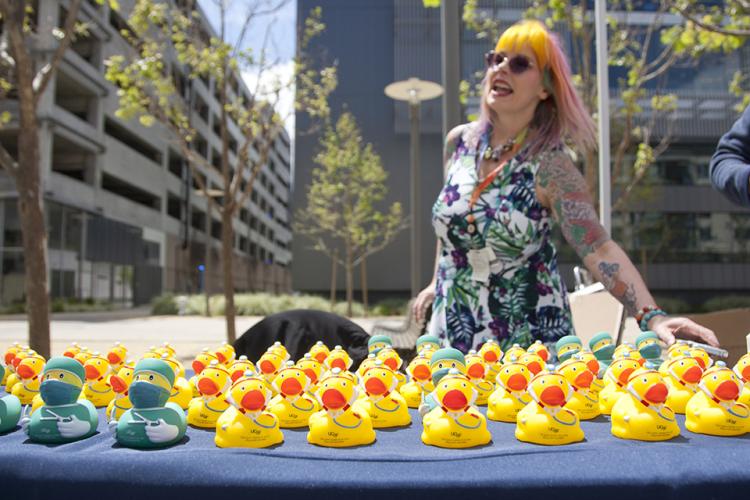 A UCSF staff member donning brightly colored hair and foliage patterned garment stands at a table outdoors with rows of yellow rubber ducks clad in medical scrubs, some with white lab coats and stethoscopes and the UCSF logo, line a table as give-away items for event attendees.