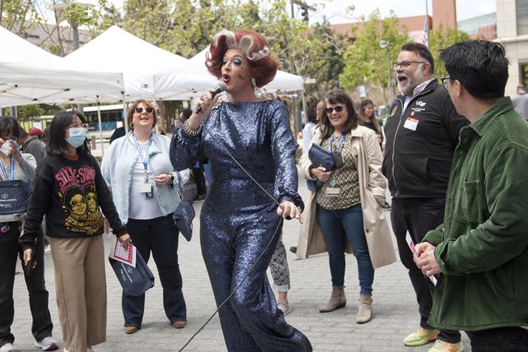 A performer in drag sings into a microphone as UCSF staff and community members gather in a semi-circle around the performance, smiling and laughing in support. 