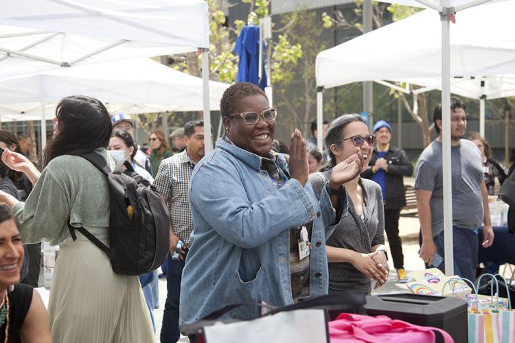 UCSF staffers, two in focus are standing at a reception table with colorful prizes. The two people in focus are laughing, smiling, and clapping as they stare off into an activity happening out of view at an outdoor event underneath white tents.