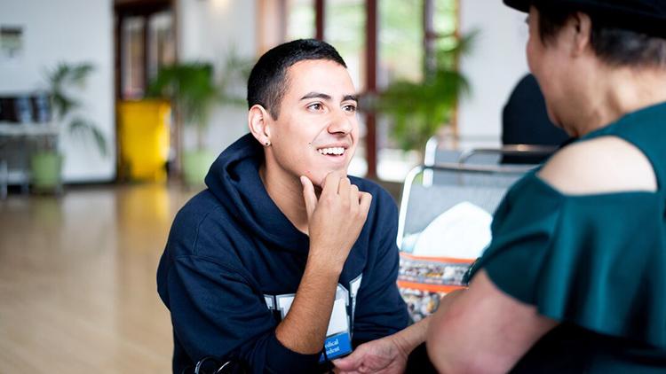 A male presenting UCSF Learner is listening and smiling, looking up towards a clinic patient in an indoor space at a free screening event hosted by the UCSF Latino Medical Student Association.