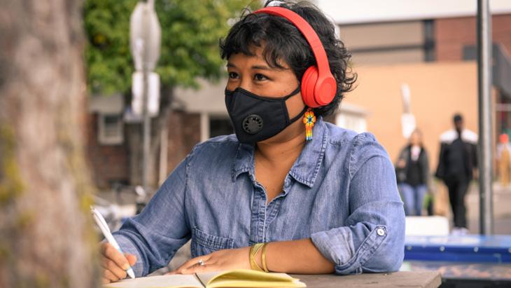 A young woman sitting at a table writing and wearing a mask and red headphones