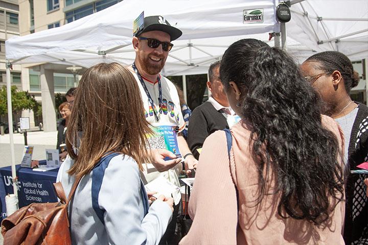 A tall, male presenting person wearing a dark baseball cap, and jersey with the UCSF Pride Pin pledges on it, handing out pins to event attendees.