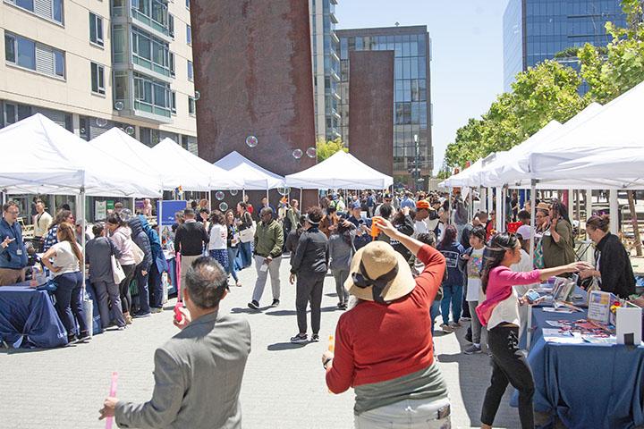 Staff Resources Day outdoor daytime event with UCSF community members visiting resource tables under white tents. Two people in the foreground have their backs to the camera while blowing bubbles.
