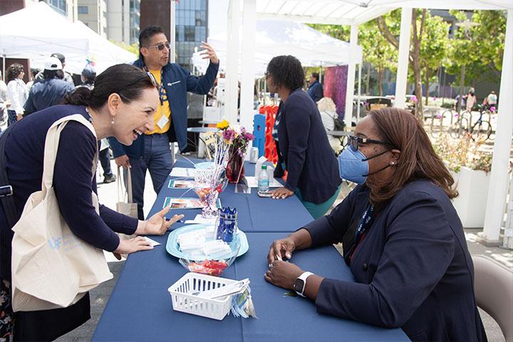 A Staff Resource Day attendee talking and smiling with a resource fair tabler who's wearing a medical facemask. Additional event participants can be seen out of focus in the background.